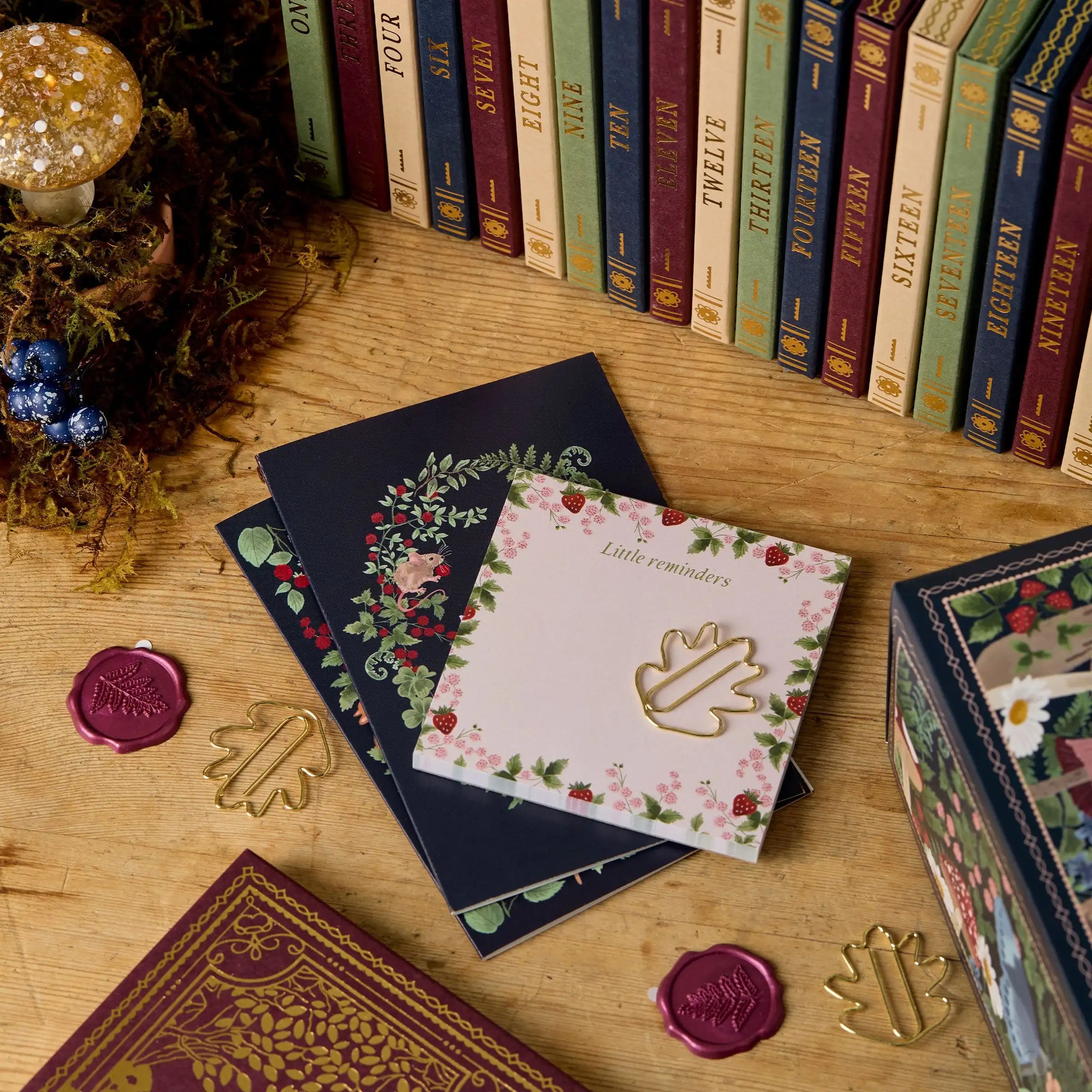 Bøger på træbord med dekorerede notesbøger og vokssegl. Vintage stil, farverige bogrygge, botaniske motiver. Perfekt til bogelskere og samlere.