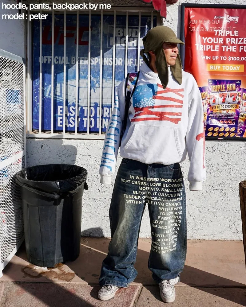 Ung mand iført hvid hættetrøje med amerikansk flag, store jeans og grøn hue foran butik. Streetwear, afslappet mode, urban stil.