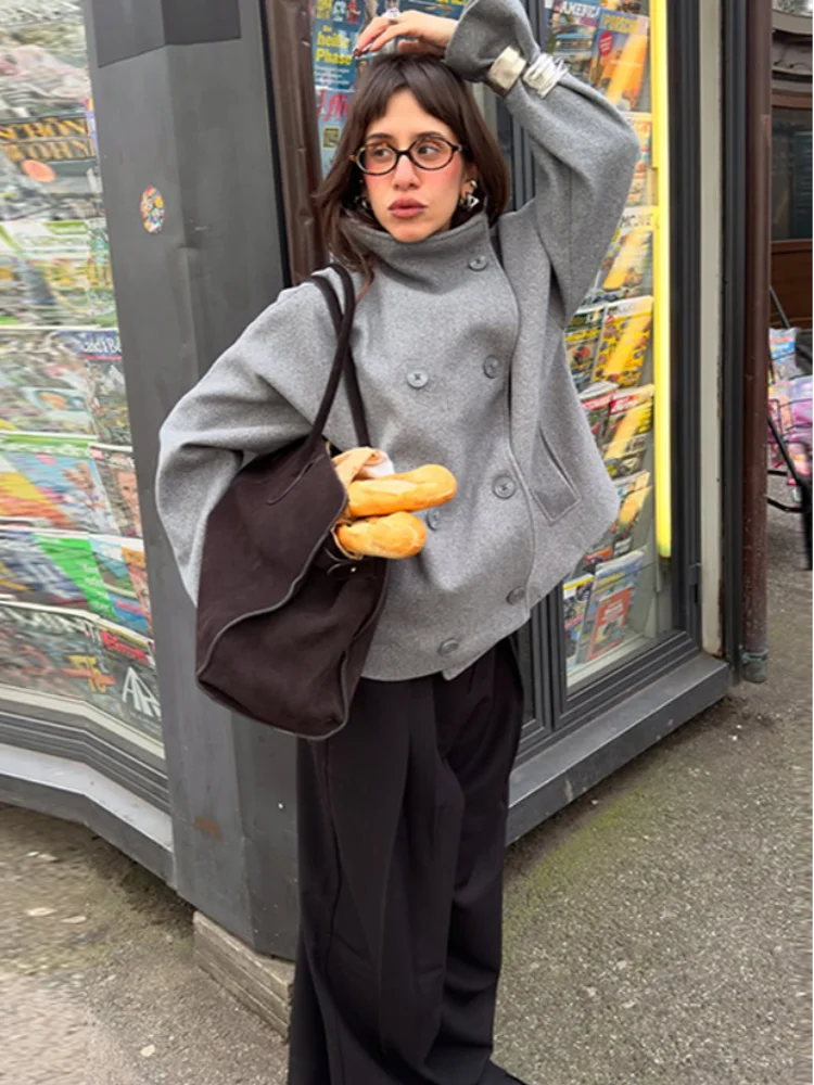 Kvinde i grå frakke med briller og sort taske, holder baguetter foran en kiosk med magasiner. Gadebillede, mode, byliv, Parisisk stil.