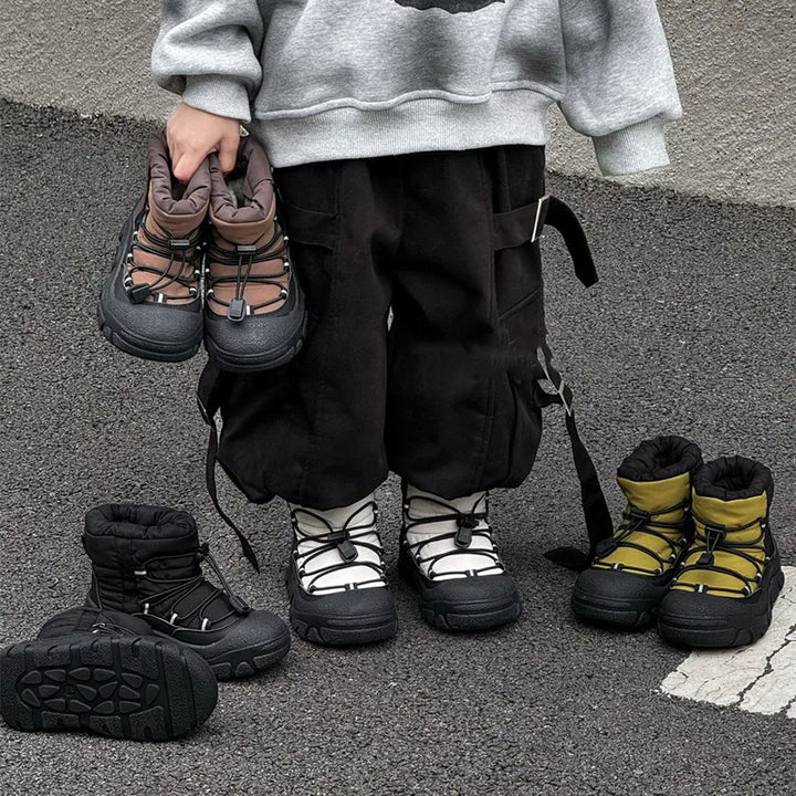 Barn iført grå sweatshirt og sorte bukser står med fire par vandrestøvler i forskellige farver på asfalt. Børnestøvler, udendørs fodtøj, vintermode.