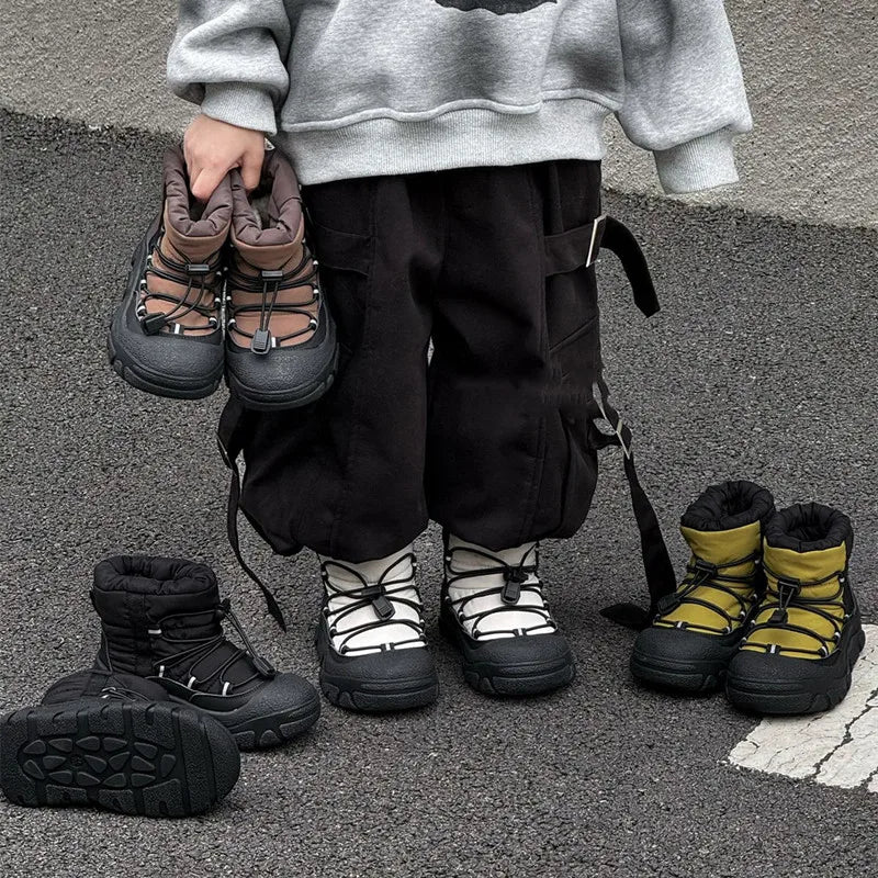 Barn iført grå sweatshirt og sorte bukser står med fire par vandrestøvler i forskellige farver på asfalt. Børnestøvler, udendørs fodtøj, vintermode.