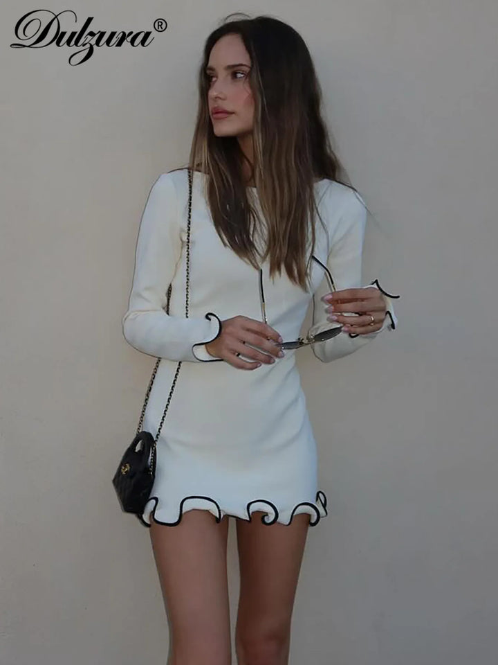 Kvinde i hvid, langærmet minikjole med sorte kanter, holder solbriller. Bærer sort skuldertaske. Mode, stilfuld, elegant kjole.