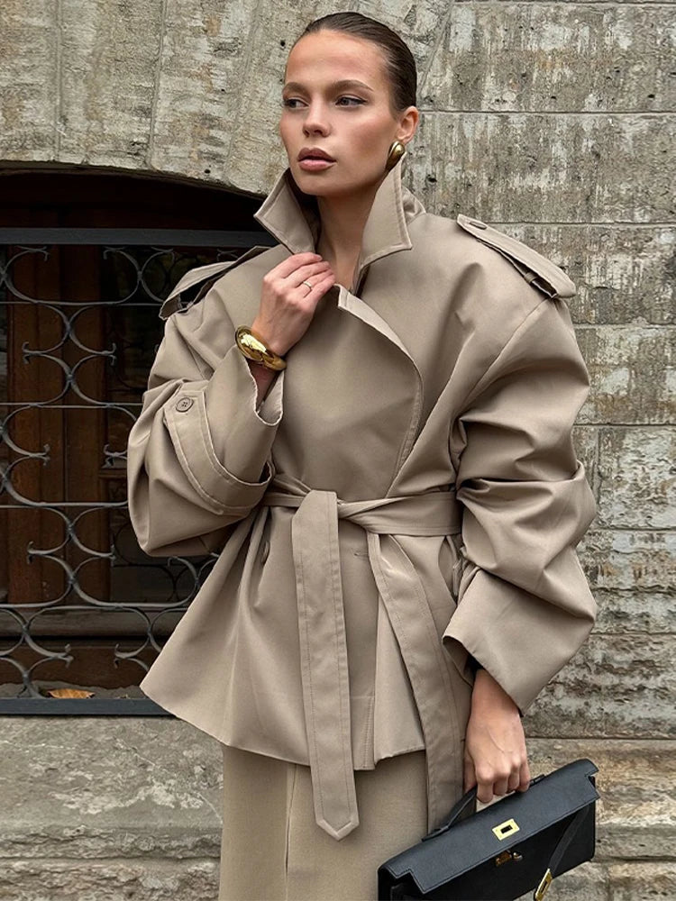 Kvinde i beige trenchcoat med bælte, holder sort taske foran stenvæg. Elegant mode, efterårsjakke, stilfuldt outfit.