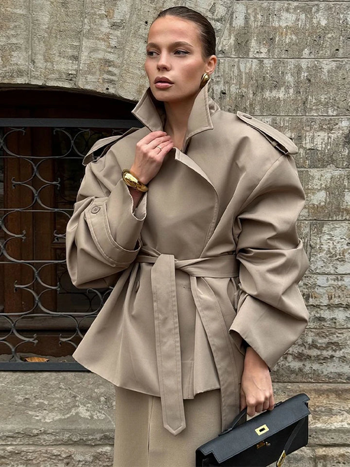 Kvinde i beige trenchcoat med bælte, holder sort taske foran stenvæg. Elegant mode, efterårsjakke, stilfuldt outfit.