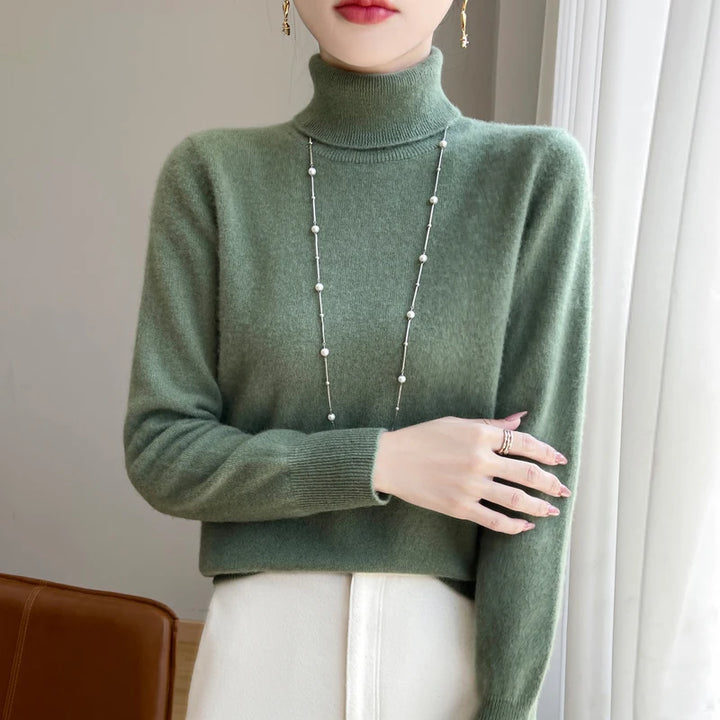 Kvinde iført grøn rullekravesweater og lang halskæde, står ved vindue. Elegant vintermode, minimalistisk stil, moderne kvindetøj.