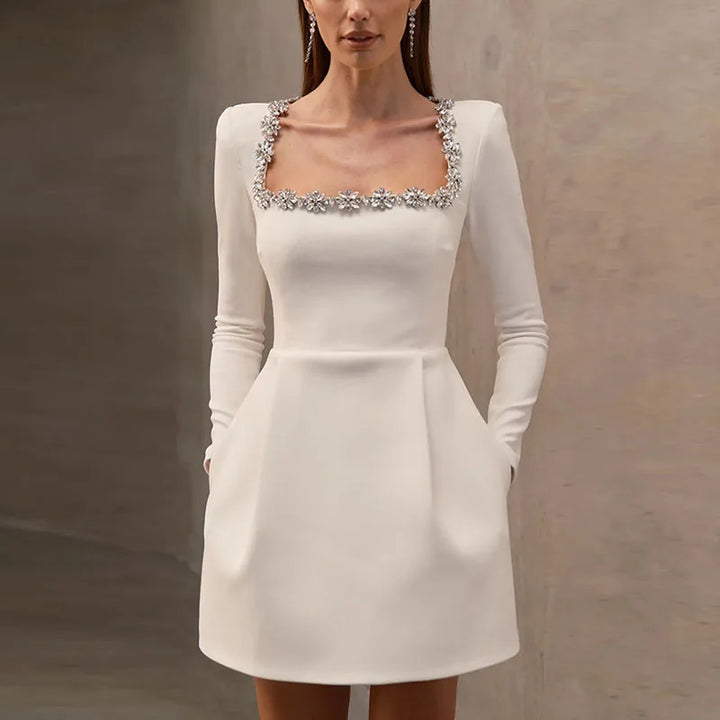 Kvinde i elegant hvid cocktailkjole med firkantet udskæring og blomsterdetaljer, lange ærmer og lommer. Perfekt til festlige lejligheder.
