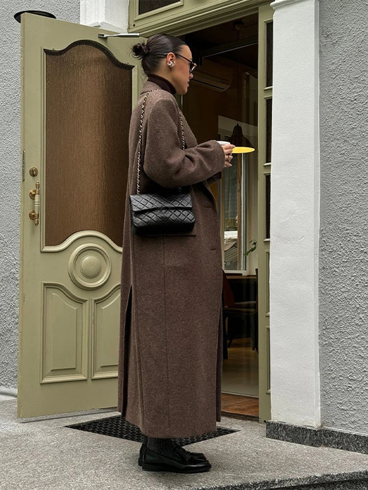 Kvinde i lang brun frakke og sort taske står ved en åben dør, iført solbriller og sorte sko. Urban mode, efterårslook, stilfuld påklædning.