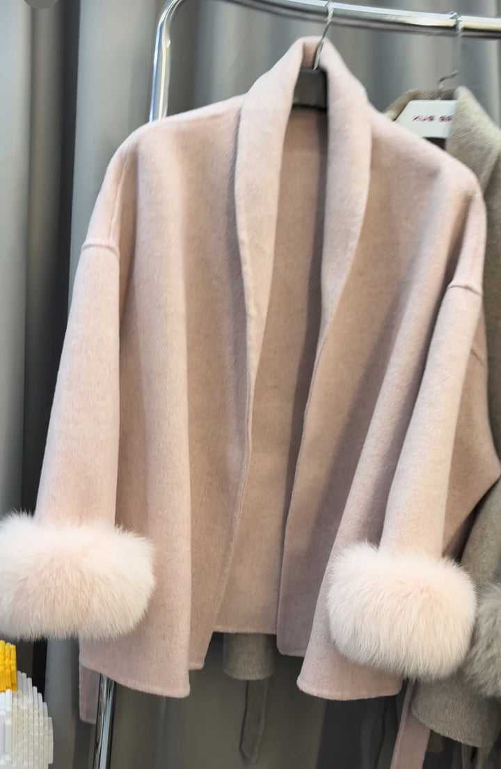 Lyserød cardigan med pelsmanchetter, hængende på tøjstativ. Blød, elegant design, perfekt til vintermode. Luksuriøs strik, trendy og stilfuld.