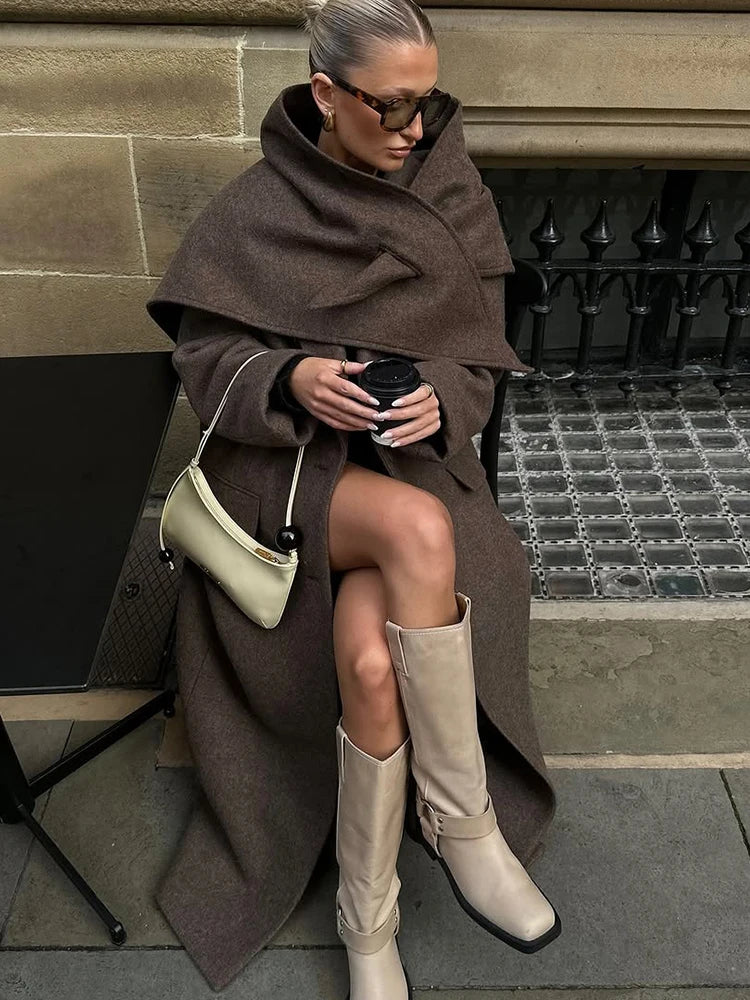 Kvinde i brunt uldfrakke og beige støvler sidder med kaffekop. Bærer solbriller og hvid håndtaske. Mode, efterår, street style.