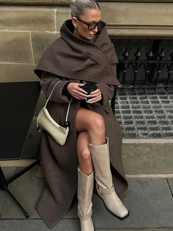 Kvinde i brunt uldfrakke og beige støvler sidder med kaffekop. Bærer solbriller og hvid håndtaske. Mode, efterår, street style.