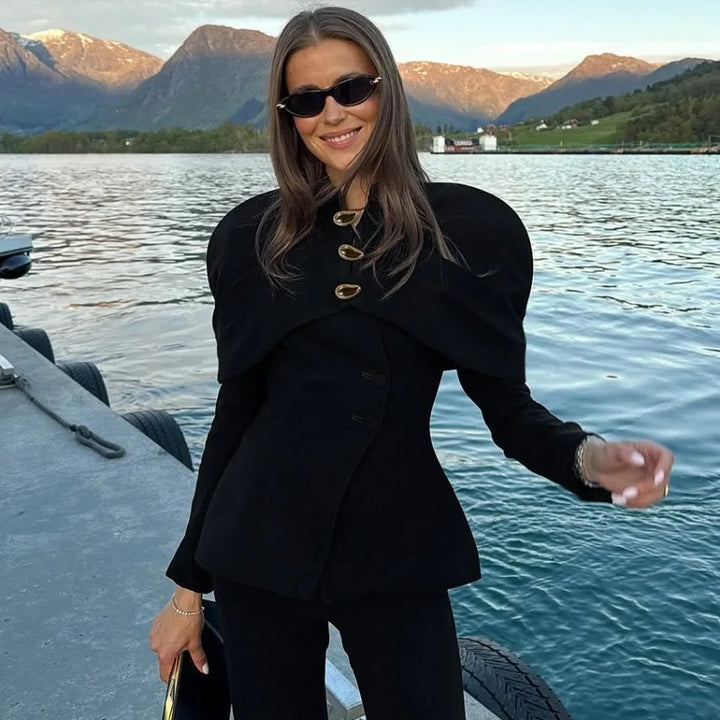 Kvinde i sort jakke og solbriller ved sø med bjerge i baggrunden. Elegant mode, udendørs, naturskøn udsigt.