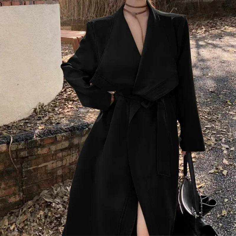 Kvinde i sort trenchcoat med bælte, står udendørs ved murstensvæg. Hun bærer en sort taske. Efterårsblade på jorden. Mode, elegant, stilfuld.