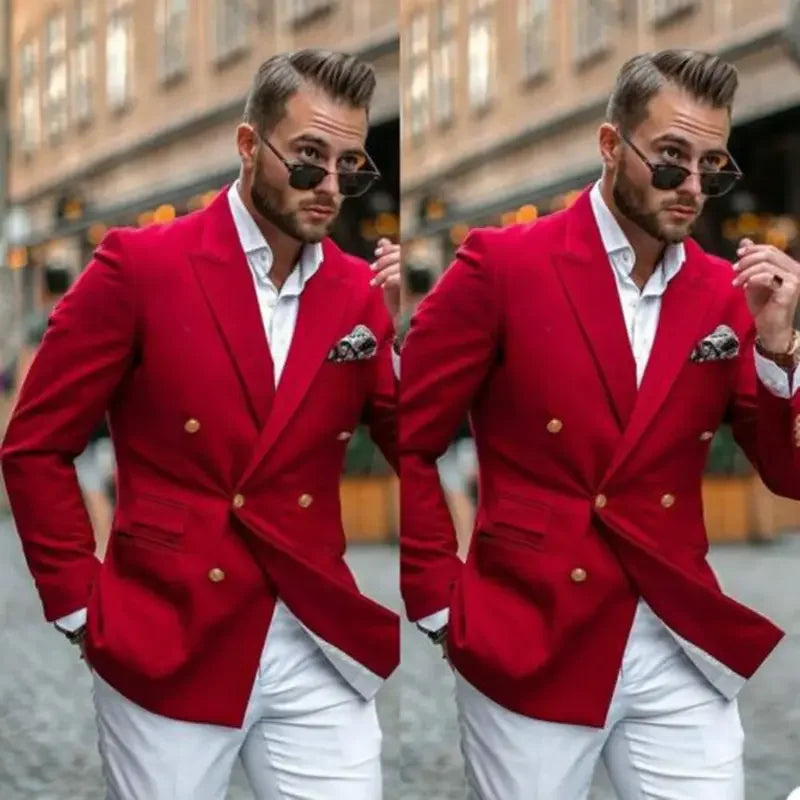 Mand i rød blazer og hvide bukser, iført solbriller, poserer selvsikkert på brostensbelagt gade. Elegant herremode, stilfuldt look.