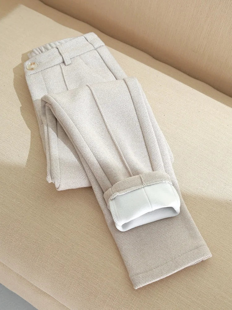 Beige bukser i herringbone-mønster, foldet på en lys sofa. Stilfuldt herretøj, moderne design, elegant og tidløst look.