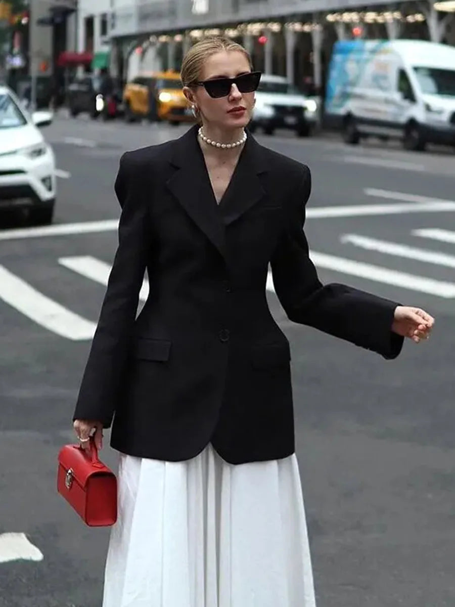 Kvinde i sort blazer og hvid nederdel på byens gade, bærer rød håndtaske og solbriller. Mode, gadebillede, stilfuldt outfit, byliv.