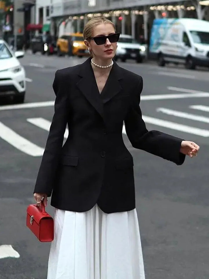 Kvinde i sort blazer og hvid nederdel på byens gade, bærer rød håndtaske og solbriller. Mode, gadebillede, stilfuldt outfit, byliv.