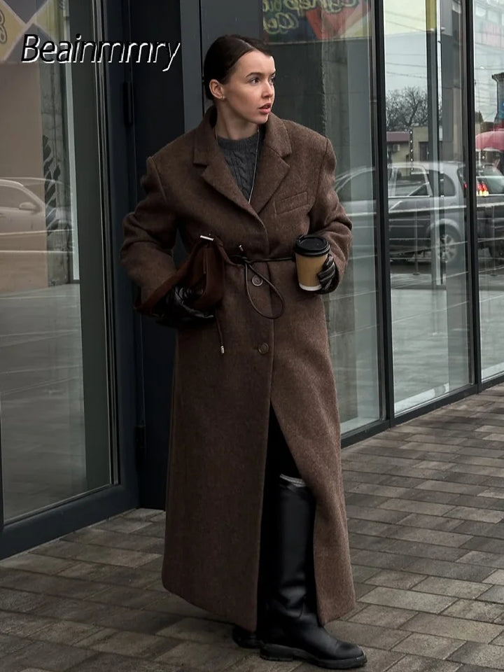 Kvinde i lang brun frakke, sorte støvler og handsker, holder en kaffekop foran en glasfacade. Vintermode, stilfuldt outfit, bymiljø.