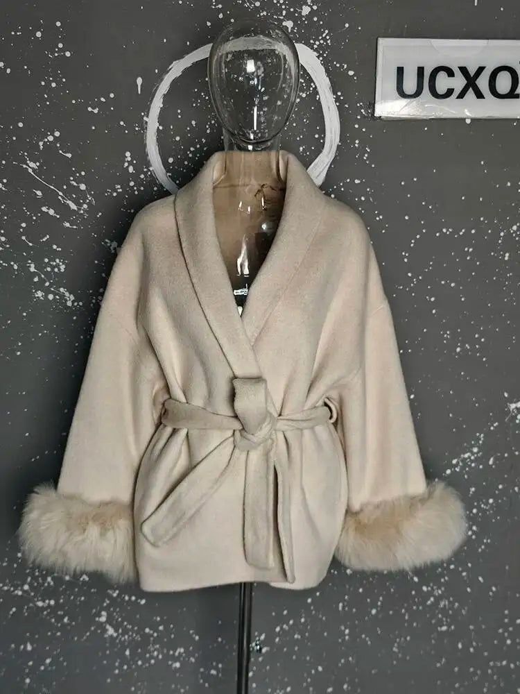 Beige frakke med bælte og pelsmanchetter på mannequin mod grå, plettet baggrund. Elegant vinterjakke, moderne design, luksuriøst tøj.