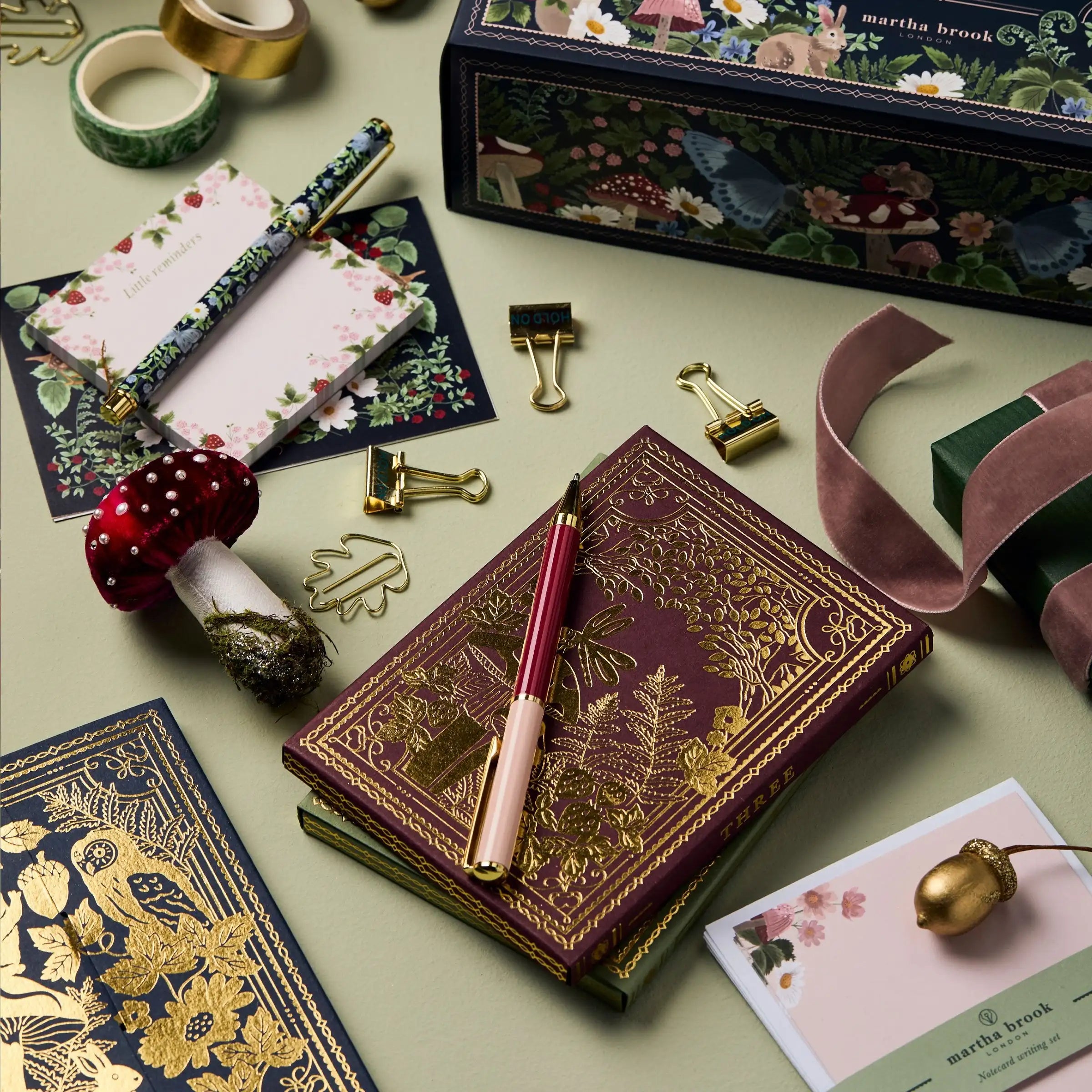 Dekorativ notesbog med guldmønster, omgivet af pen, svampepynt, klips og gaveæske. Perfekt til bullet journaling og kreativ skrivning.