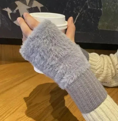 Person iført grå, fluffy fingerløse handsker holder en hvid kaffekop på et træbord. Vintermode, varm drik, hyggelig stemning.