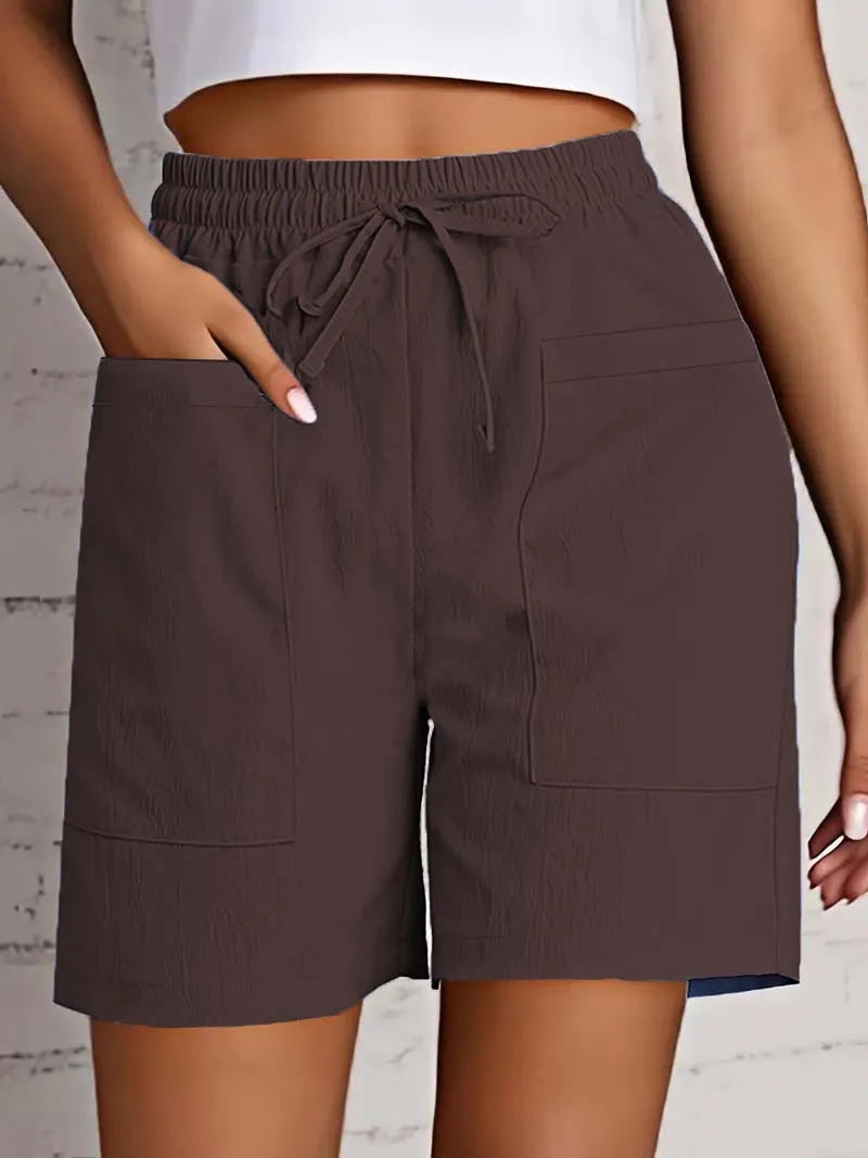 Kvinde iført brune shorts med elastisk talje og snøre, store lommer, mod hvid murstensvæg. Komfortabelt sommertøj, casual stil.