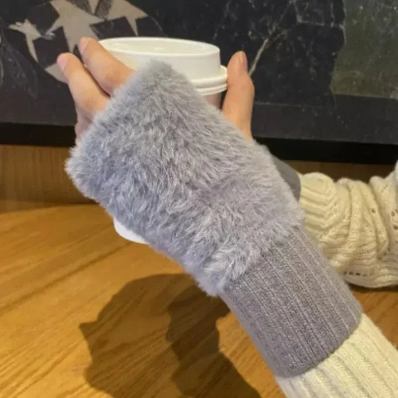 Hænder iført grå, fluffy fingerløse handsker holder en hvid kaffekop. Varmt vintertilbehør, hyggelig caféstemning, strikket sweater.