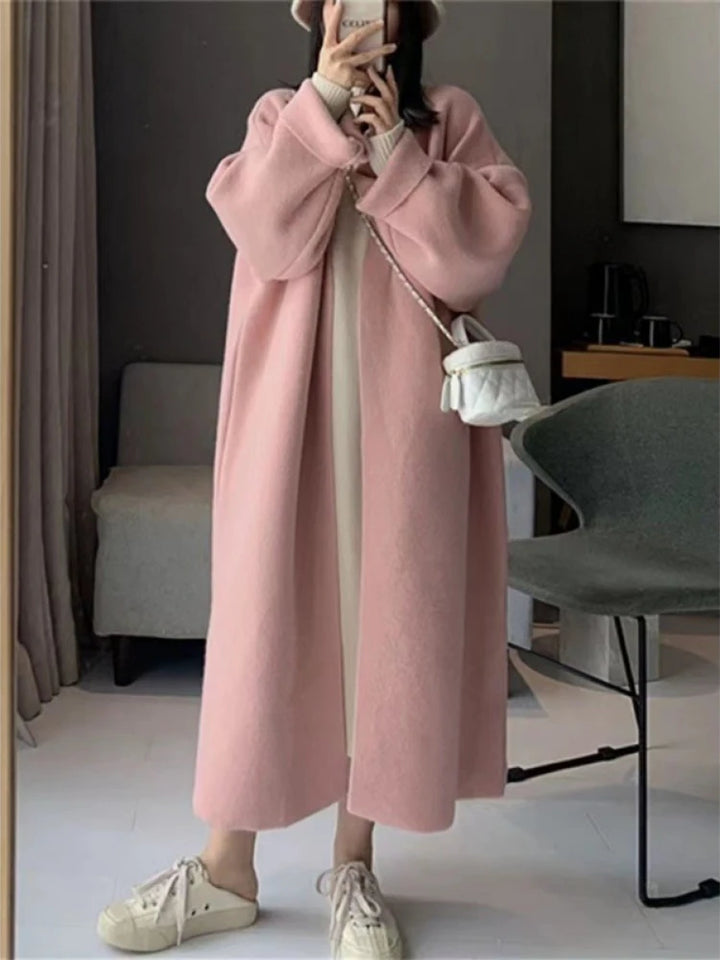 Kvinde iført lang lyserød frakke, hvid kjole og sneakers, poserer indendørs med hvid skuldertaske. Moderne mode, vintertøj, stilfuldt look.