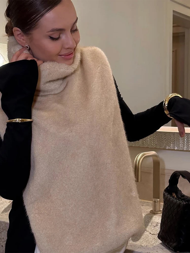 Kvinde iført beige strikponcho og sort trøje, smilende ved køkkenvask. Elegant mode, vintertøj, moderne stil, gyldne armbånd.