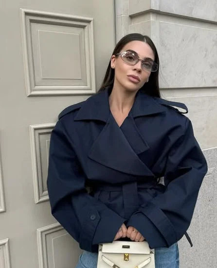 Kvinde i mørkeblå trenchcoat og solbriller, står foran en dør, holder en hvid taske. Mode, stil, efterårstøj, moderne kvindemode.