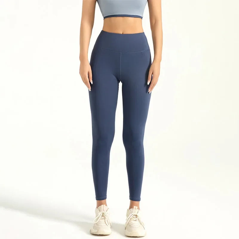 Kvinde iført blå leggings og grå sports-bh, står på hvid baggrund. Komfortabelt træningstøj, perfekt til fitness og yoga.