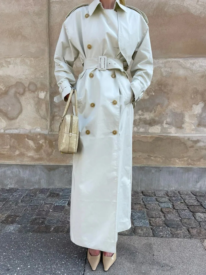 Dame i lang, beige trenchcoat med bælte og guldknapper, holder en flettet taske. Står på brostensbelagt gade foran en murstensvæg.