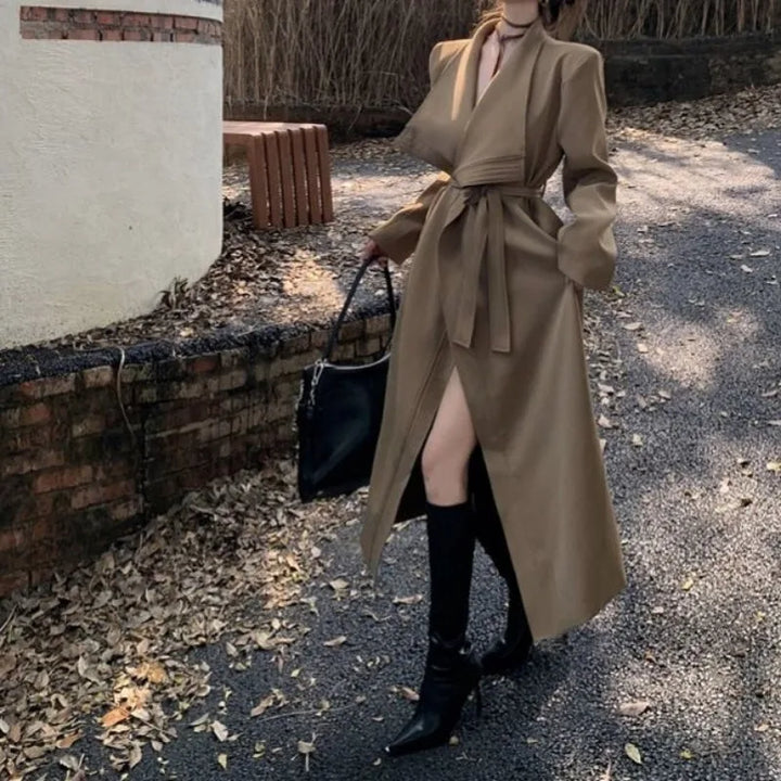 Kvinde i lang brun frakke med bælte, sorte støvler og taske, går på bladstrøet sti. Elegant efterårsoutfit, mode, udendørs stil.