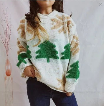 Kvinde iført striktrøje med grønne juletræsmotiver og beige mønstre, stående foran en hvid baggrund. Vintermode, hyggelig sweater, casual stil.