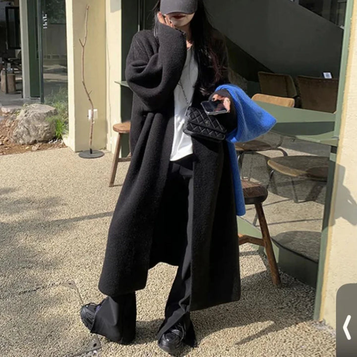 Kvinde i lang sort frakke og sorte bukser, holder en blå jakke og sort quiltet taske. Står udenfor café med glasvægge. Mode, efterår, street style.