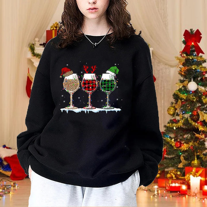 Ung kvinde i sort sweatshirt med julemotiv af tre vinglas, står foran juletræ. Perfekt til julemode, vintertøj, festlig stil.