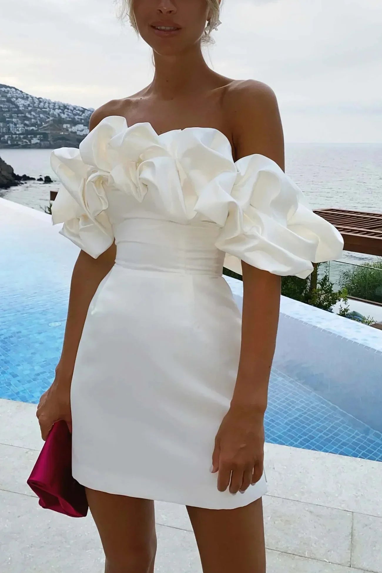 Kvinde i elegant hvid cocktailkjole med voluminøse ærmer ved pool med havudsigt. Perfekt til sommerfest eller bryllup.