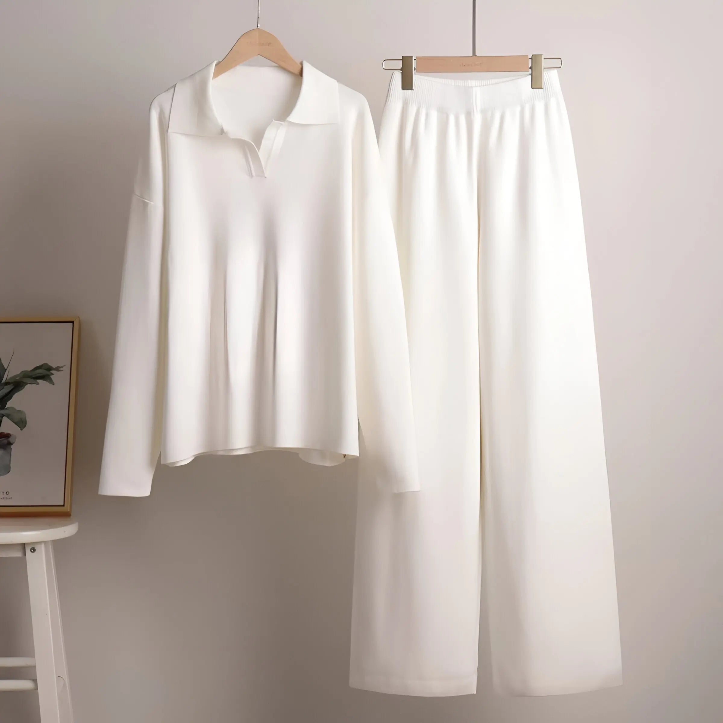 Hvidt sæt tøj med langærmet bluse og brede bukser hængende på bøjler. Minimalistisk design, moderne stil, perfekt til casual eller formel brug.