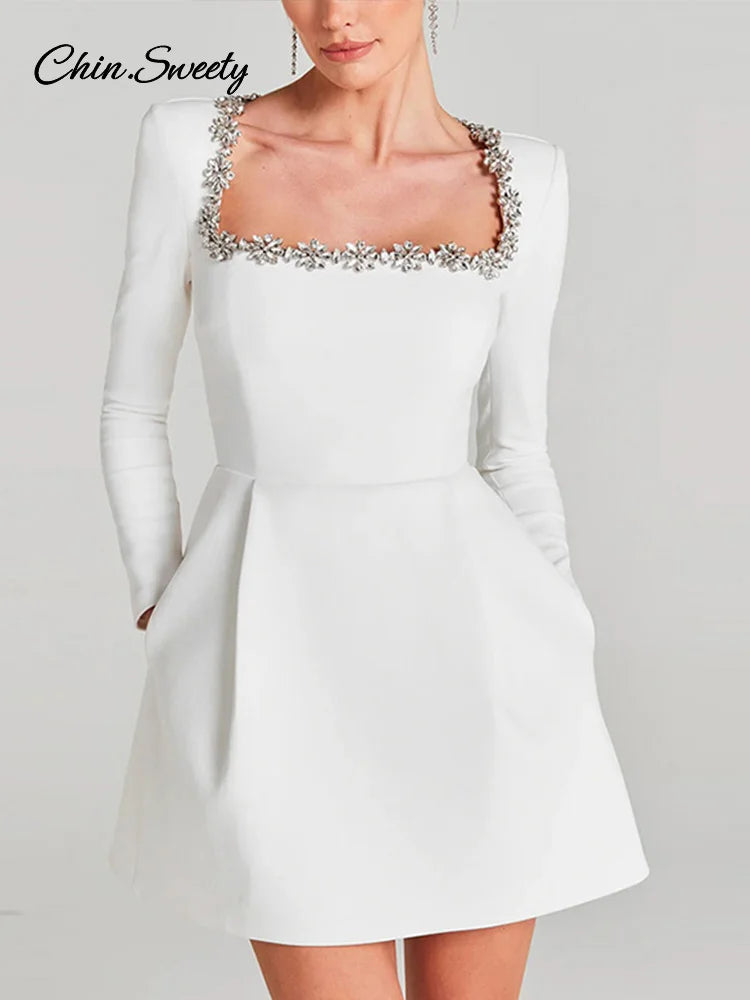 Hvid cocktailkjole med lange ærmer og firkantet halsudskæring dekoreret med blomsterdetaljer. Elegant og stilfuldt design til festlige lejligheder.