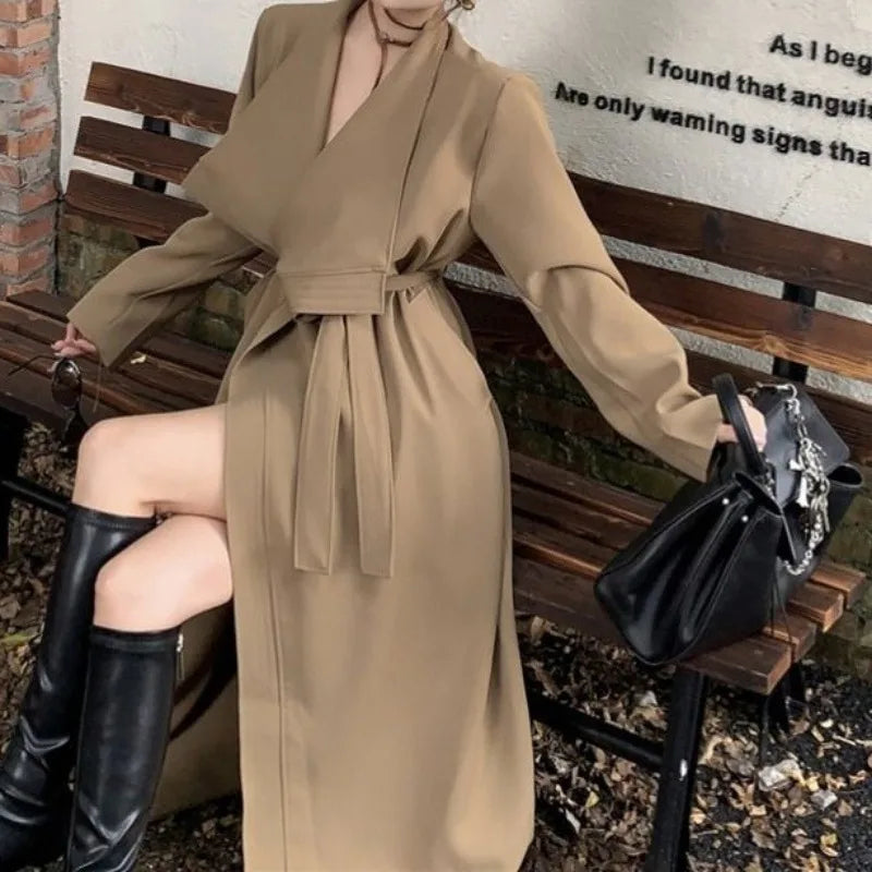 Kvinde i beige trenchcoat sidder på bænk, iført sorte knæhøje støvler og holder en sort taske. Elegant mode, efterårsoutfit, stilfuldt look.