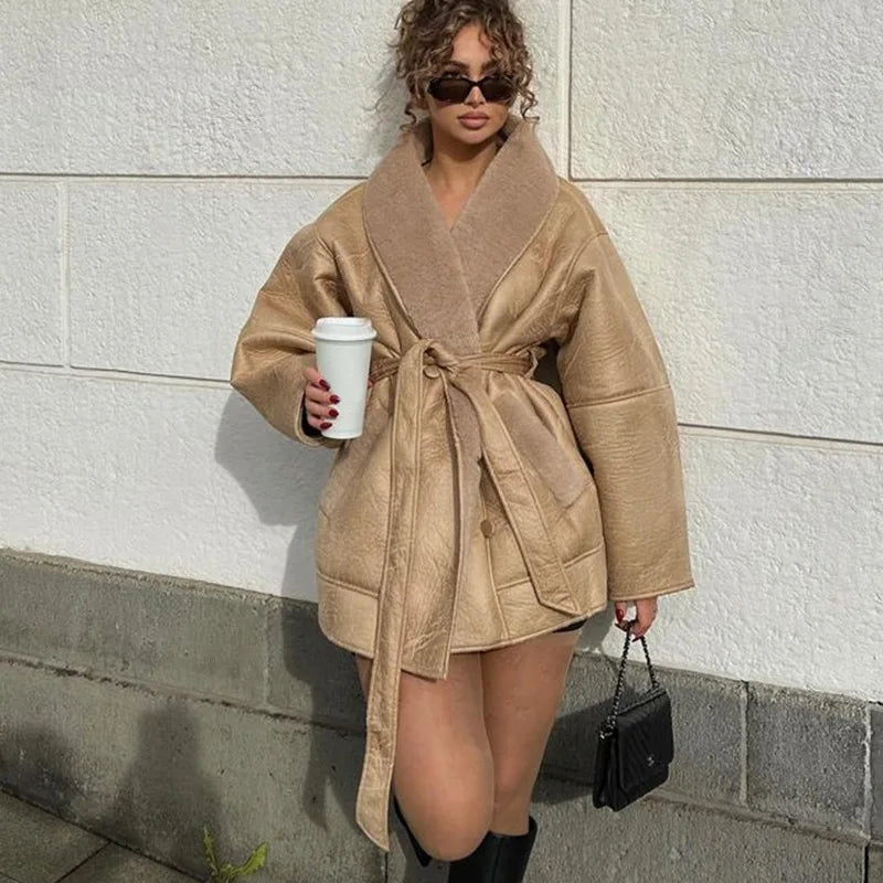 Kvinde iført beige frakke med bælte, solbriller og sorte støvler, holder en hvid kaffekop og sort taske, står mod hvid væg. Trendy vintermode.