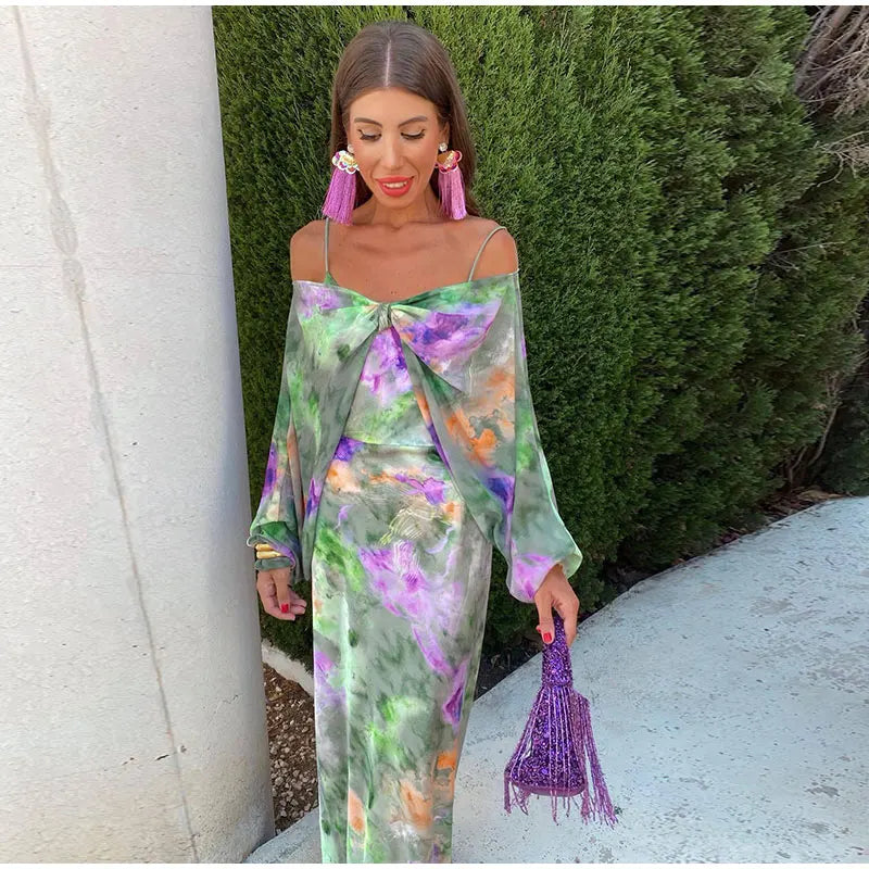 Kvindelig model i farverig, blomsterprintet kjole med lilla frynseøreringe og matchende taske, poserer udendørs ved grøn busk.