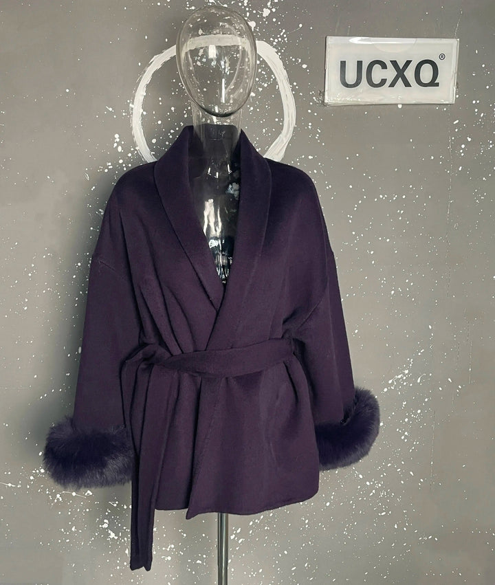 Lilla kåbe med bælte og pelsmanchetter på mannequin, UCXQ logo i baggrunden. Elegant vintermode, stilfuldt design, varm beklædning.