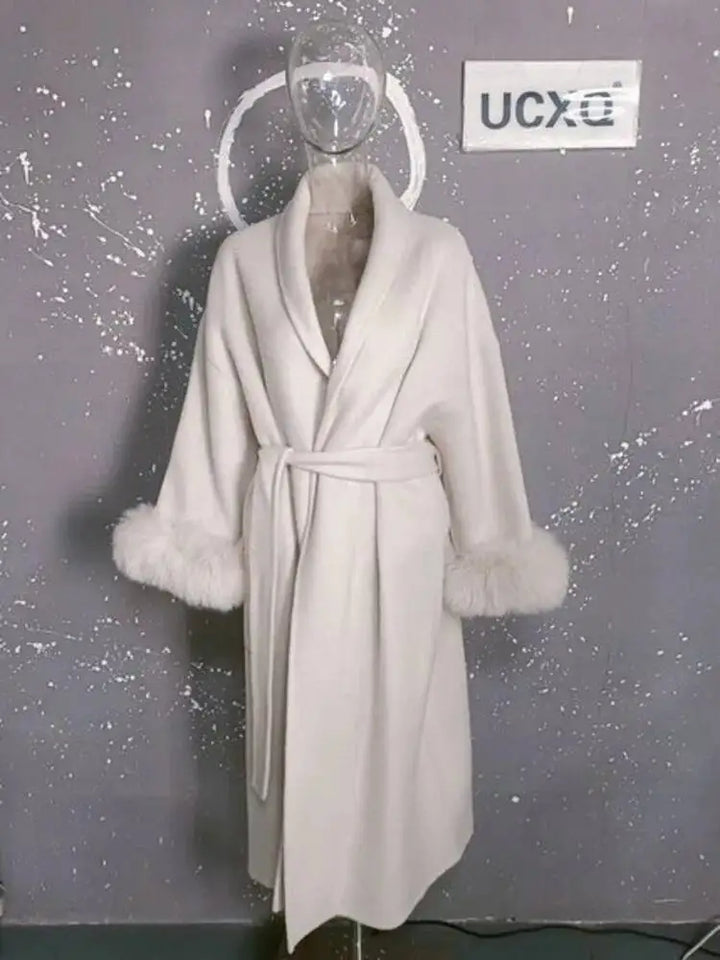 Hvid frakke med pelsmanchetter på gennemsigtig mannequin mod grå baggrund. Elegant vintermode, luksuriøs damefrakke, stilfuldt design.