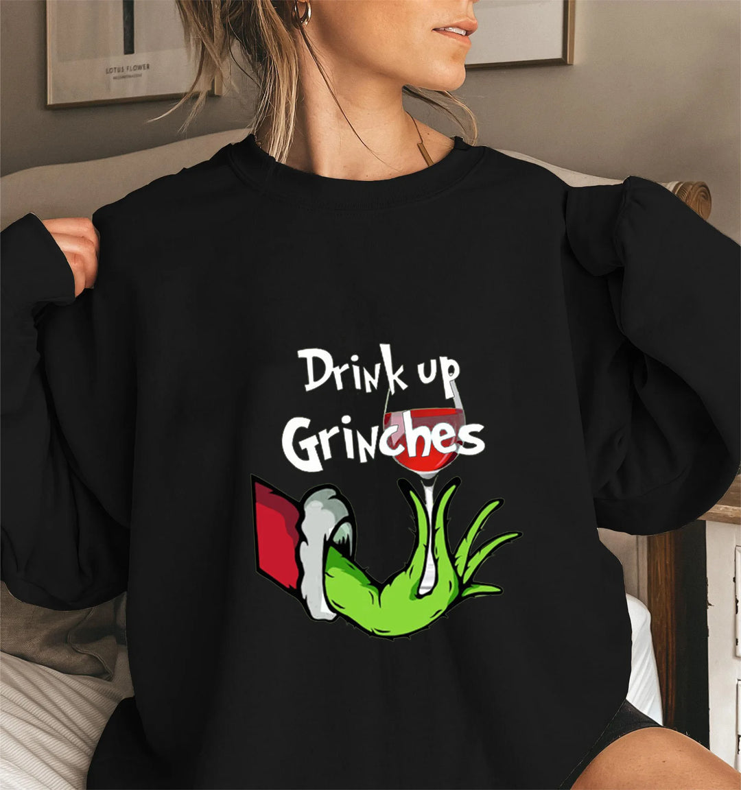 Kvinde iført sort sweatshirt med "Drink Up Grinches" tekst og grøn hånd, der holder et glas vin. Sjovt julemotiv, perfekt til festlig påklædning.