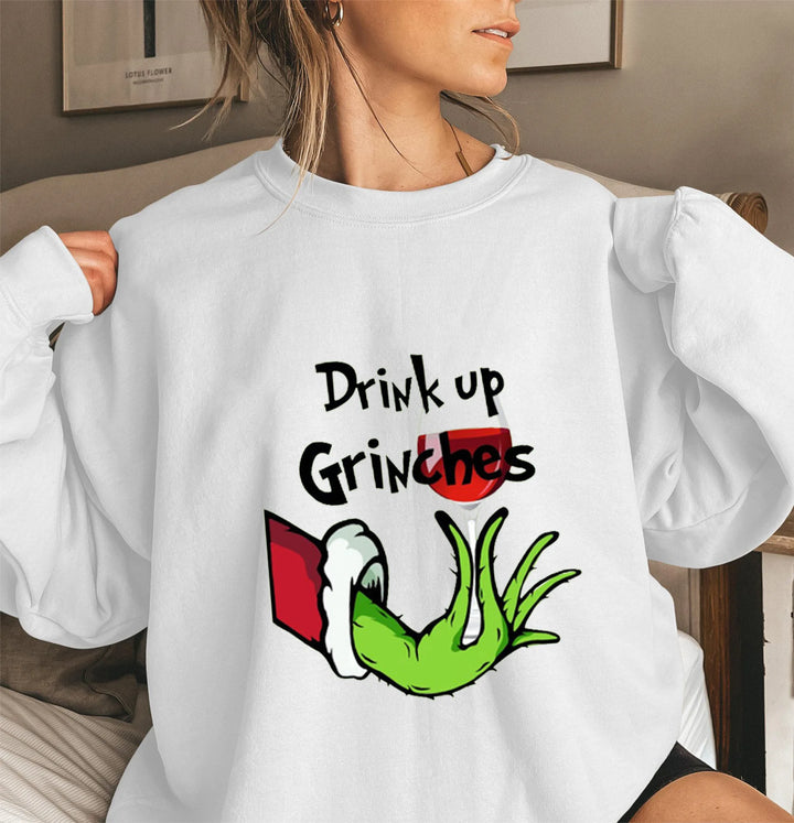 Kvinde iført hvid sweatshirt med "Drink up Grinches" og en grøn hånd i julemotiv. Perfekt til jul, sjovt tøj, vintermode.
