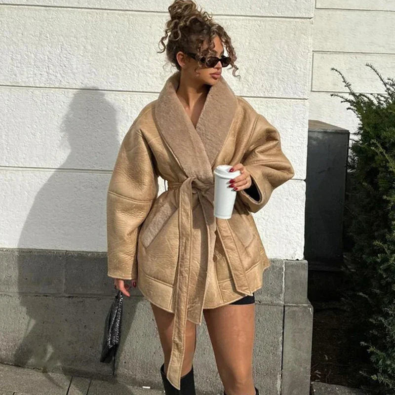 Kvinde iført beige, oversized frakke med bælte, solbriller og krøllet hår, holder en hvid kop. Mode, vinterjakke, street style, udendørs.