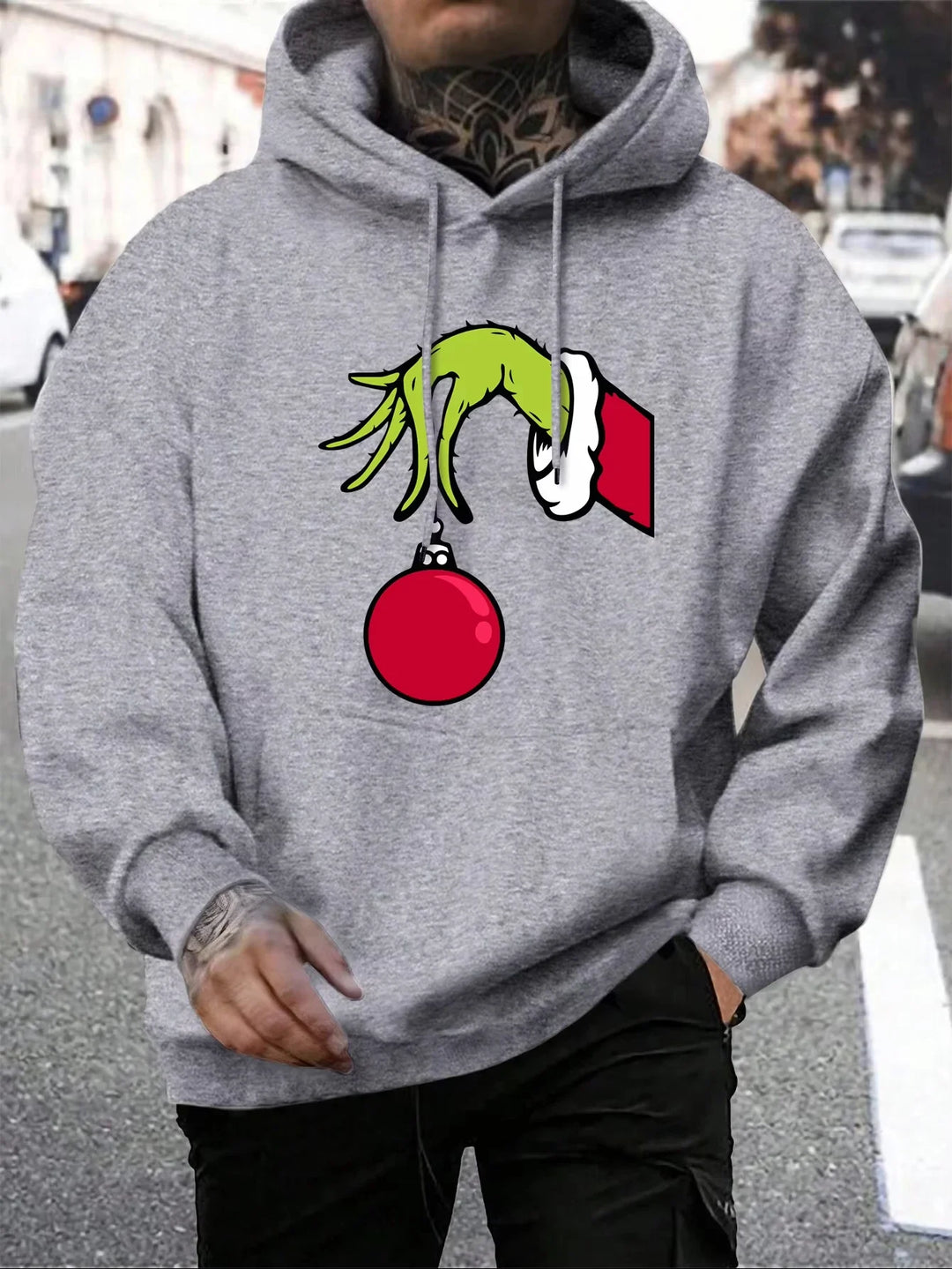 Grå hættetrøje med Grinch-hånd, der holder en rød julekugle. Perfekt til jul, unisex design, streetwear stil.