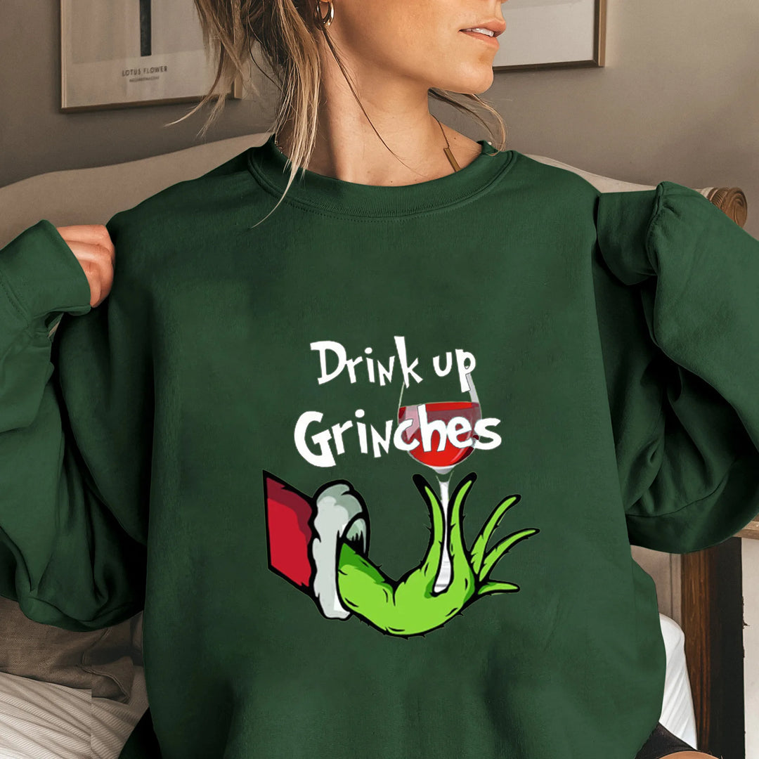 Kvinde iført grøn sweatshirt med "Drink up Grinches" tekst og tegning af grøn hånd, der holder et glas vin. Perfekt til julefester og sjove outfits.