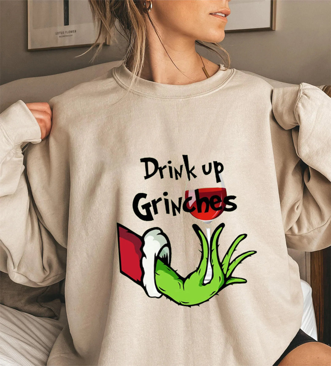 Kvinde iført beige sweatshirt med Grinch-motiv og teksten "Drink up Grinches". Perfekt til jul, sjovt design, populær juletrøje.
