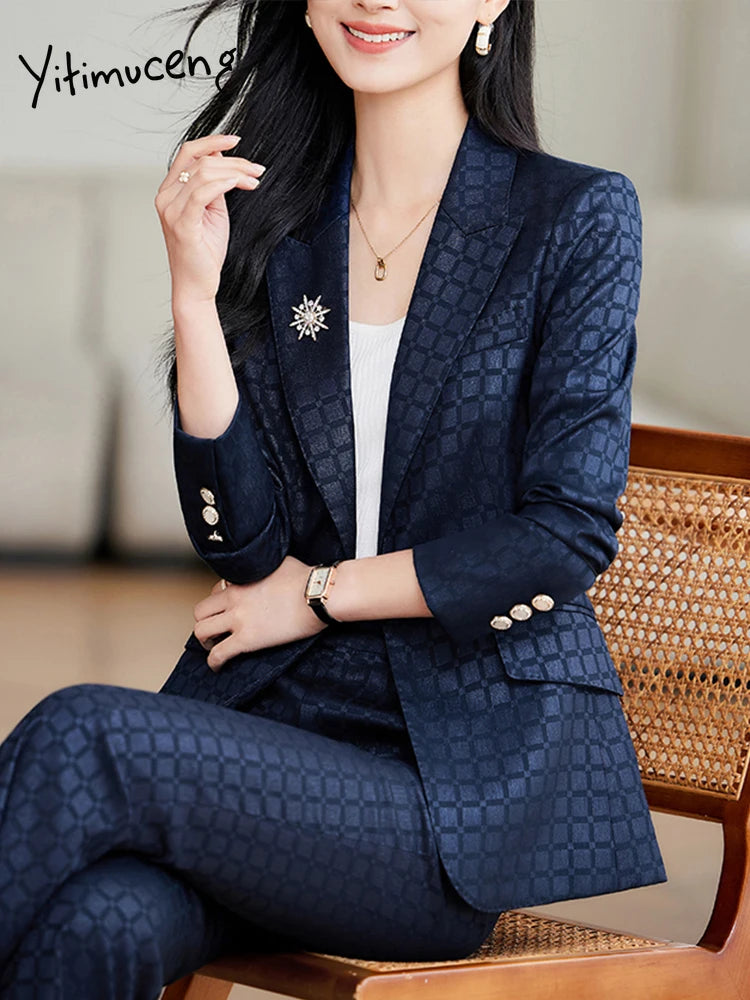 Kvinde i elegant, mørkeblå jakkesæt med mønster, sidder på stol. Bærer hvid top, smykker og broche. Moderne businessmode, stilfuldt look.